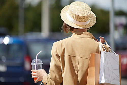 Rear view of fashionable young woman in straw hat and beige jacket holding shopping bags and takeout cup of juice and leaving mall