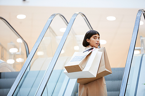 Portrait of beautiful young woman holding shopping bags with Black Friday looking over shoulder at camera while going up escalator in mall, copy space