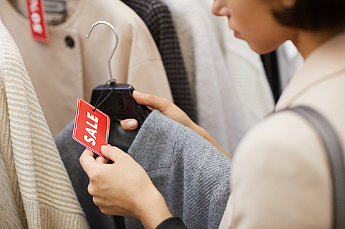 Closeup of unrecognizable young woman holding autumn coat with red SALE tag while browsing clothes in shopping mall on Black Friday, copy space