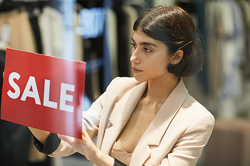 Portrait of beautiful woman hanging red SALE sign on window display in clothes store, copy space
