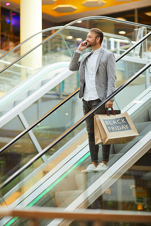 Portrait of fashionable man holding shopping bags with Black Friday inscription while speaking by phone standing on escalator in mall, copy space