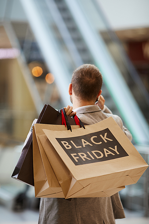 Back view portrait of fashionable man holding shopping bags with Black Friday inscription while speaking by phone standing against escalator in mall, copy space