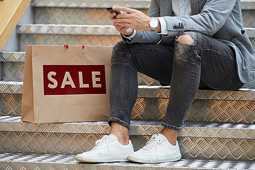 Closeup of fashionable young man using smartphone sitting on stairs in mall, shopping bags with SALE inscription beside him, copy space
