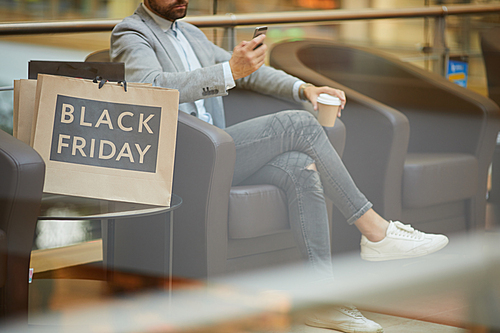 Cropped portrait of fashionable young man using smartphone sitting in comfortable armchair in mall, shopping bags with Black Friday inscription beside him, copy space