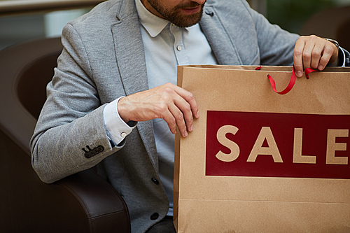 Mid-section portrait of fashionable man holding paper bag with red SALE inscription while shopping in mall, copy space