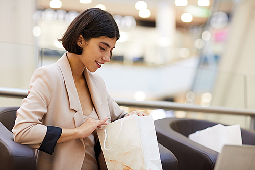 Portrait of beautiful young woman looking into paper bag and smiling happily while enjoying shopping spree in mall, copy space