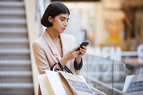 Waist up portrait of elegant young woman holding shopping bags and using smartphone while standing in mall on Black Friday, copy space