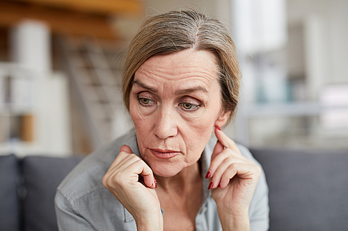 Portrait of sad mature woman sitting on couch at home and looking away with worry and anxiety, copy space