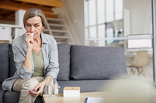 Portrait of sad mature woman sitting on couch at home and crying holding tissue, copy space