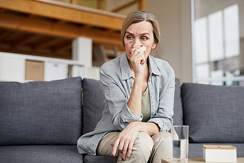 Portrait of crying senior woman sitting on couch at home and holding tissue, copy space