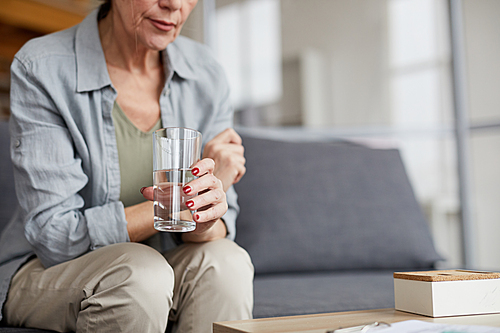 Cropped portrait of sad senior woman holding glass of water sitting on couch while trying to calm down, copy space