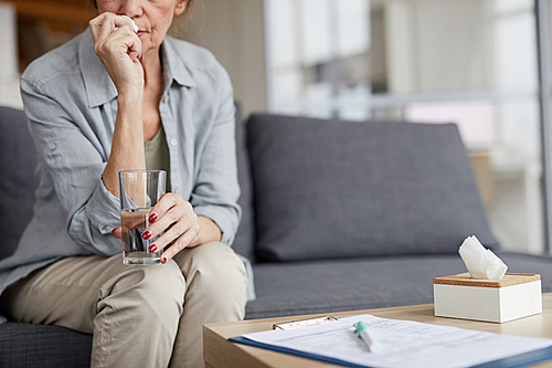 Cropped portrait of crying senior woman holding glass of water sitting on couch while trying to calm down, copy space