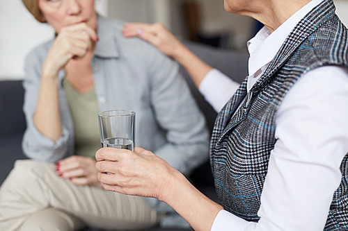 Cropped image of female psychologist comforting patient during therapy session and offering glass of water, copy space