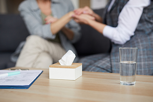 Closeup of female psychologist holding hands of senior patient while comforting her during therapy session, focus on tissue box in foreground, copy space