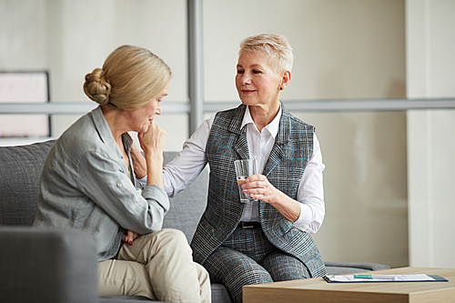 Portrait of caring female psychologist offering glass of water to crying senior woman while comforting her during therapy session, copy space