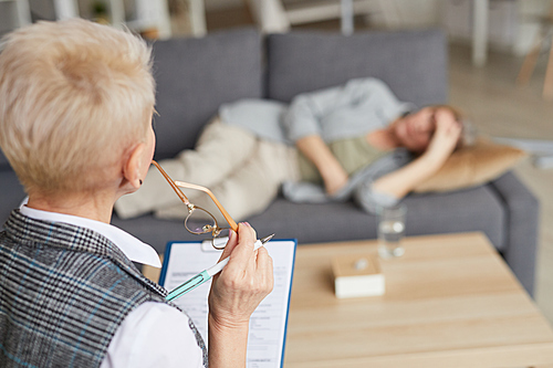 High angle portrait of female psychologist filling in medical form while listening to patient lying on couch during therapy session, copy space