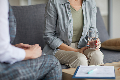 Cropped portrait of adult woman holding glass of water while sharing troubles with psychologist during therapy session, copy space