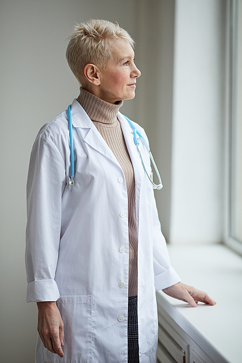 Waist up portrait of mature female doctor standing by window in clinic and looking away pensively
