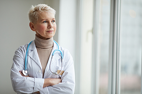 Waist up portrait of mature female doctor standing with arms crossed by window in clinic and looking away pensively, copy space