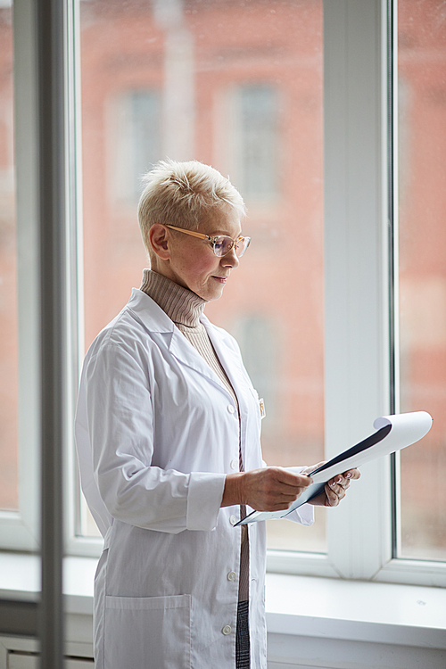 Side view portrait of mature female doctor holding clipboard while standing by window in clinic and reading patient forms, copy space