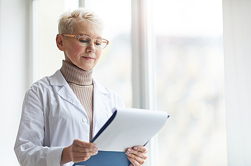 Waist up portrait of mature female doctor holding clipboard while standing by window in clinic and reading patient forms, copy space