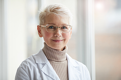 Head and shoulders portrait of friendly female doctor smiling at camera while standing against window in clinic, copy space