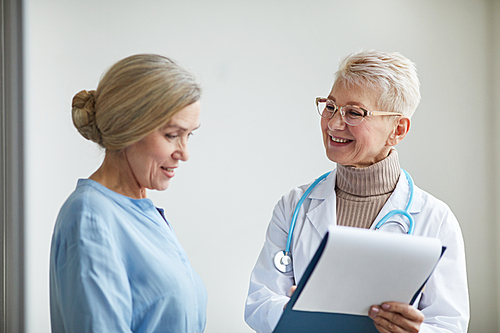Waist up portrait of mature female doctor smiling cheerfully while talking to senior woman preparing her for medical procedures in clinic, copy space