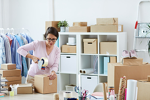 Young brunette woman sealing box with online order of client with cellotape while working in office