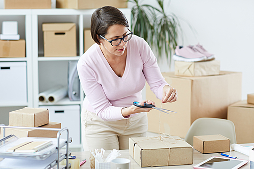 Young brunette manager of online shop cutting tied thread on top of box after packing order of client