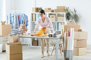 Young manager standing by desk while folding yellow pullover before packing it and sending to client