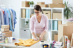 Young casual female bending over desk while folding cotton pullover before putting it into box