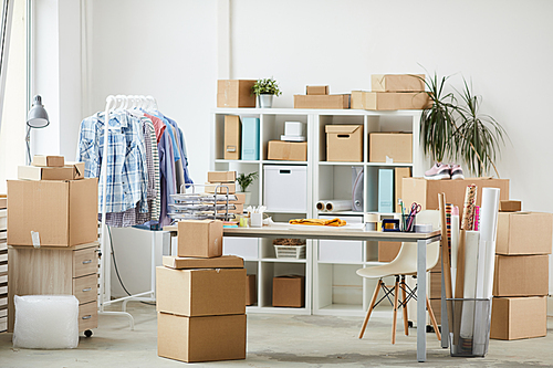 Group of supplies on desk, stacks of boxes, rack with casualwear and shelves in office of contemporary online shop
