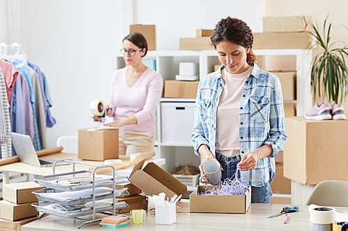 Pretty young manager of online shop packing mug in carton box on background of colleague