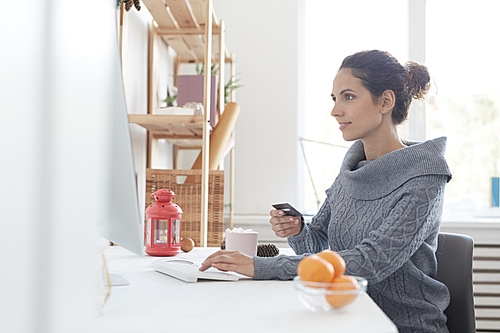 Positive beautiful young woman in gray sweater sitting at table with decorations and using computer while paying online with credit card