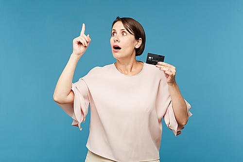 Amazed woman with black credit card showing gesture with pointed forefinger on blue background