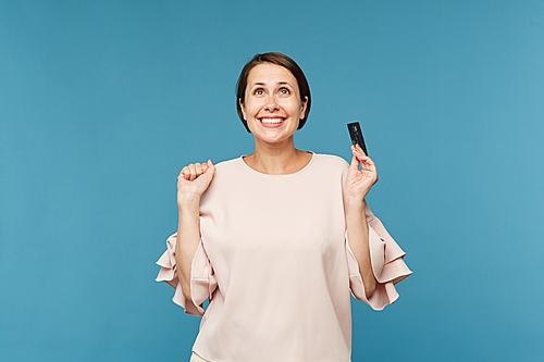 Young cheerful woman with credit card expressing joy while looking upwards over blue background in isolation