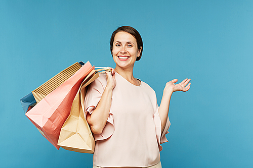 Young satisfied shopaholic with several paperbags standing in front of camera on blue background