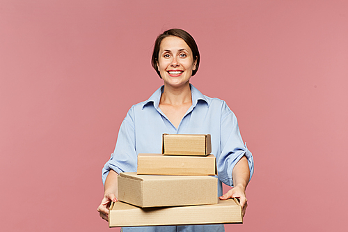 Young cheerful female online shopper with stack of packed orders looking at you on pink background