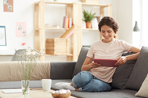 Happy pretty young woman in casual outfit sitting on sofa at home and using tablet while browsing internet