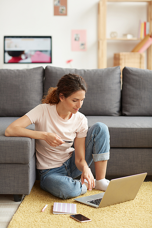 Content attractive young woman in casual outfit sitting on carpet in living room and using laptop while paying via e-commerce website