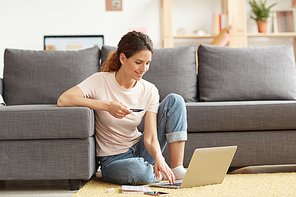 Attractive young woman sitting on carpet at home and using laptop while doing shopping on internet