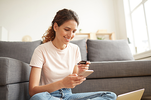 Smiling beautiful girl with ponytail sitting on floor and using smartphone while paying for online course with credit card