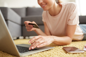 Close-up of smiling young woman lying on carpet and using credit card and laptop while doing online shopping at home