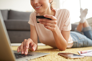 Close-up of happy girl lying on floor at home and checking card details on laptop while making online order