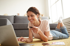 Jolly beautiful young lady in casual clothing lying in relaxed pose on floor and using laptop while paying online with credit card