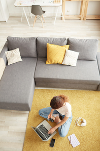 Above view of young woman with curly hair sitting with crossed legs on carpet and using laptop while paying for online order