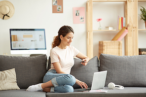 Cheerful modern girl in tshirt and jeans sitting on sofa at home and using debit card while buying goods in online store