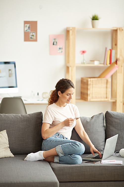 Content beautiful young woman in jeans sitting on comfortable sofa in living room and using credit card to make online payment on website