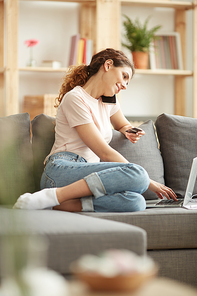 Smiling young woman with curly hair sitting on sofa and inserting number of credit card for purchasing while talking on mobile phone