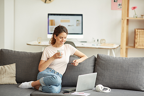 Positive attractive young woman sitting on comfortable sofa and drinking coffee while doing online shopping